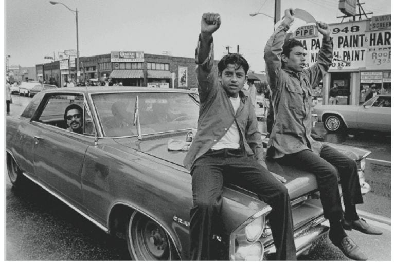 Two young Chicano men ride on the hood of a car during a national Chicano Moratorium Committee march in opposition to the war in Vietnam.