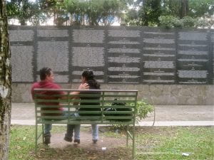 A couple sit on a bench across from a memorial wall.