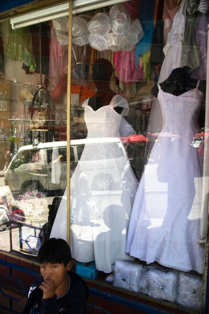Photo of a young man sitting on a bench in front of a bridal shop, with two wedding dresses in the window display.