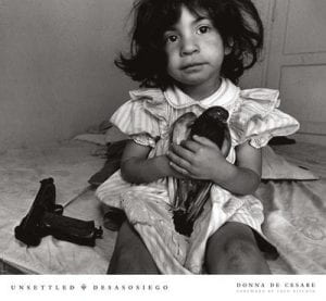 Cover of book, Unsettled/Desasosiego, Children in a World of Gangs, by Donna DeCesare. The cover photo shows a young girl sitting on a bed with a bird in her hand and a handgun next to her.