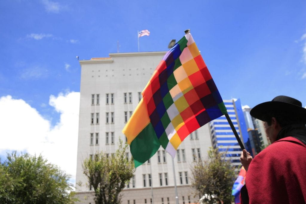 In front of the U.S. Embassy, an indigenous man waves the Bolivian Wiphala flag.