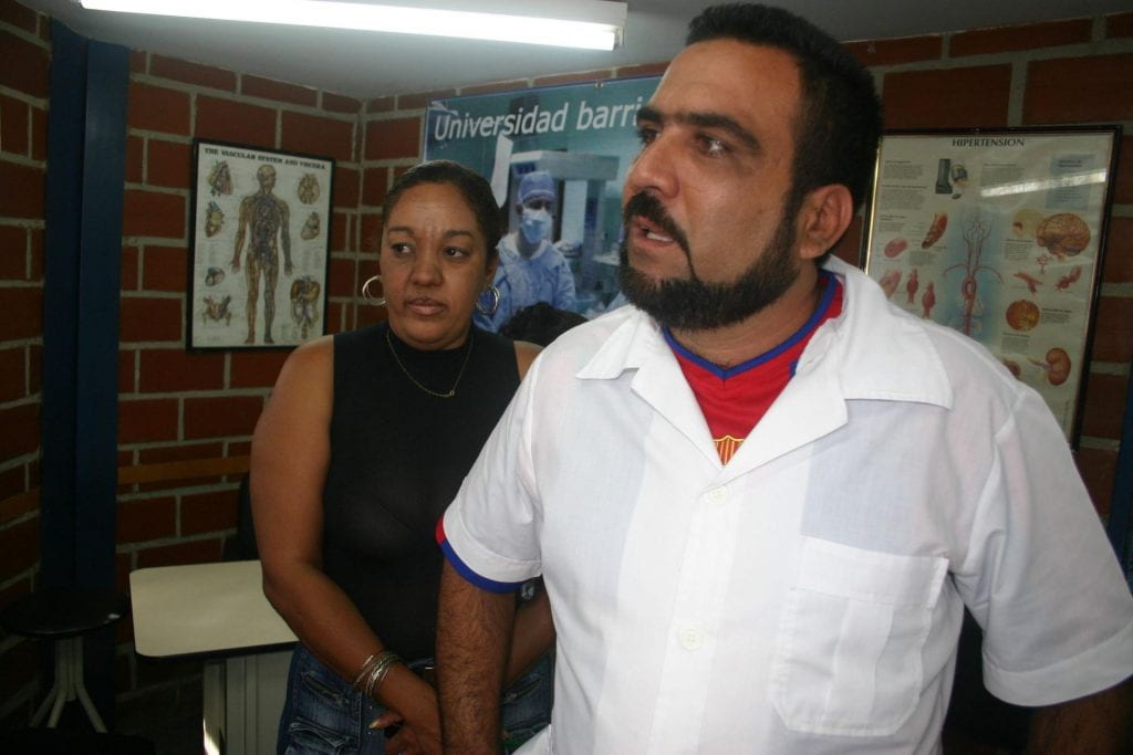 A male Cuban doctor with beard, wearing a white short-sleeve shirt and red undershirt, stands in a brick office, which has several medical posters on the wall.