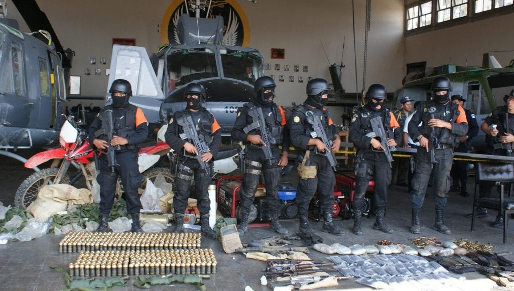 A photo showing an arsenal of weapons and drugs seized during a raid. Behind the weapons and drugs stand six police with machine guns.