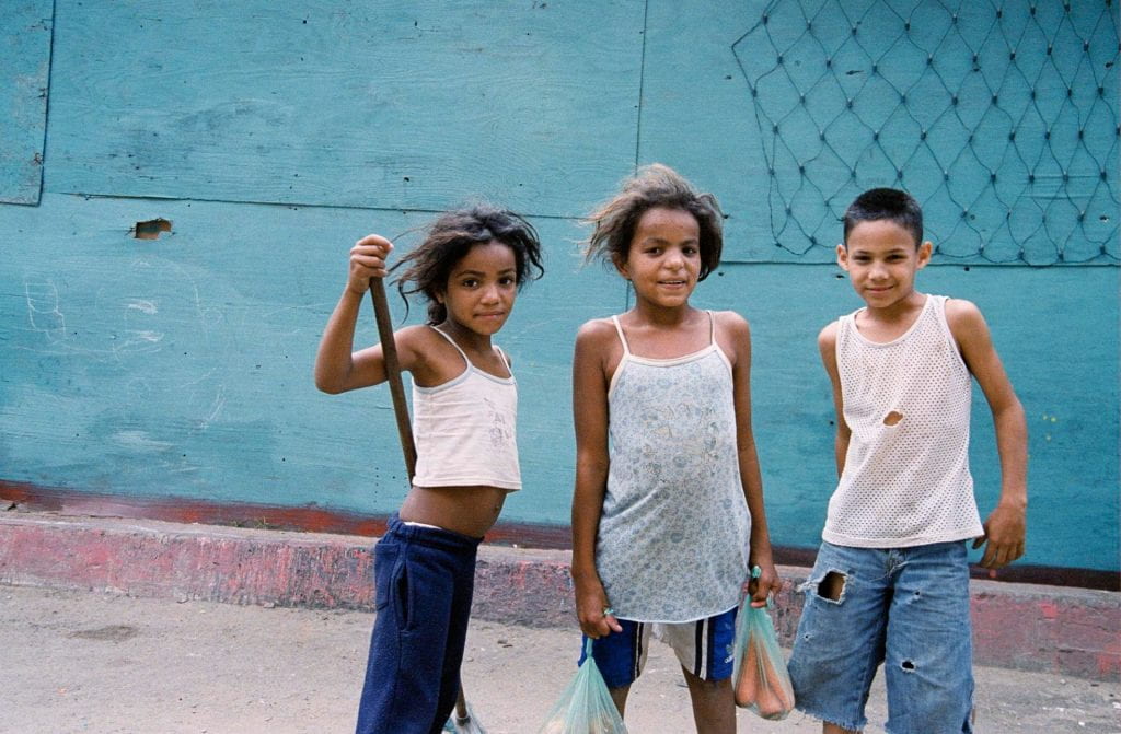 Three school-age children pose for a picture. One of them is carry plastic bags of vegetables.