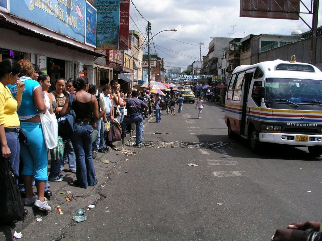 On a city street, people wait in a long line. Lines for buses and food supplies are a common sight.