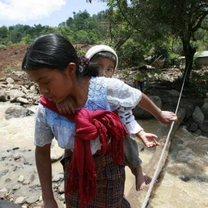Photo of a mother carrying a child on her back, crossing a stream by walking over a tree trucnk. A rope has been attached to a tree to guide her.