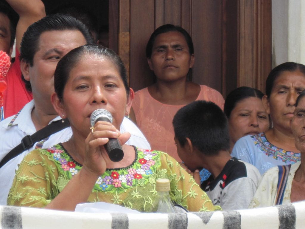 María Maquín, with microphone in hand, speaks out against the military base at a rally, surrounded by people.