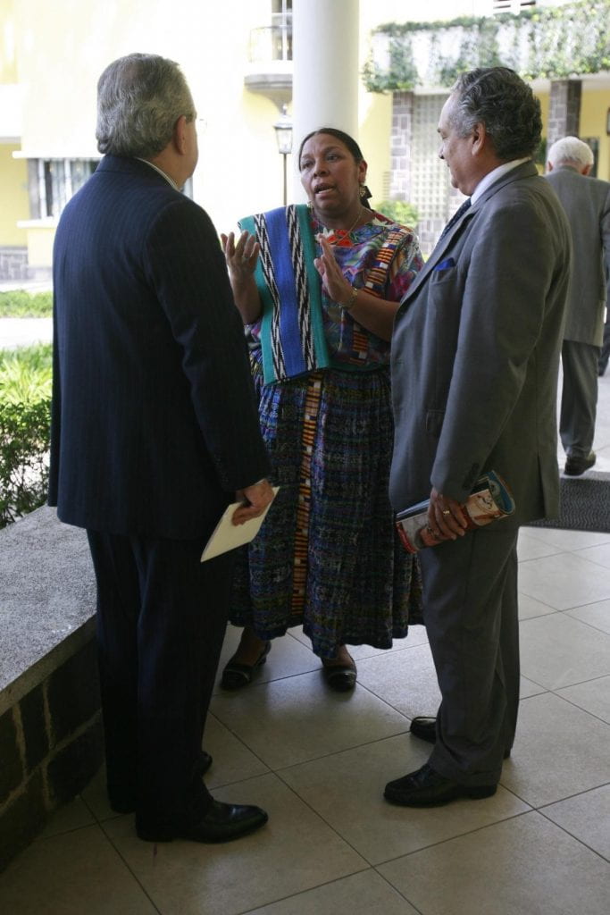 A professional Mayan woman wearing professional indigenous clothing engages in a conversation with two men in western business suits.