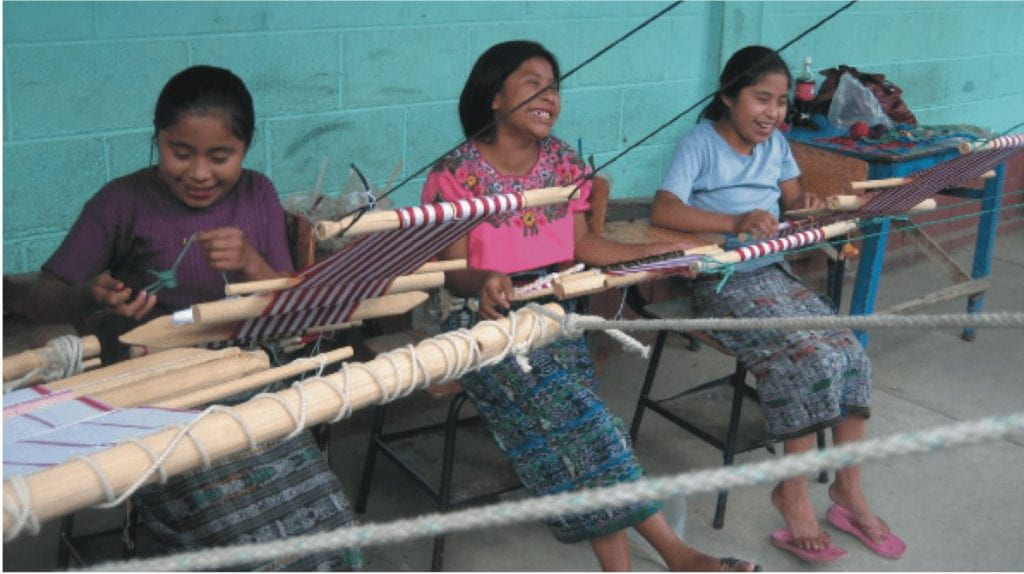 Photo of three indigenous girls at weaving tables.