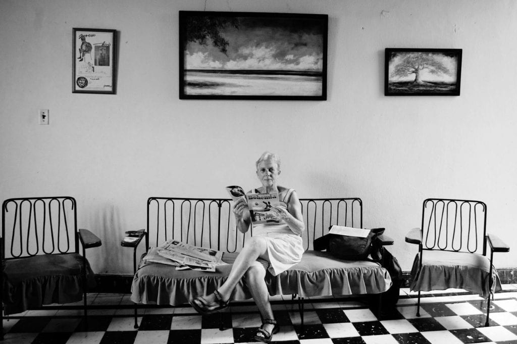 Photo of Dulce, a female writer and party activist in Matanzas, Cuba. She sits on s small sofa, with three paintings hanging on the wall behind her, and a checkerboard floor.