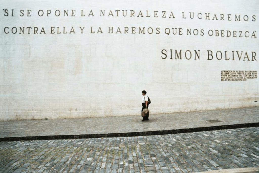 A person walks by a large stone wall, with the letters displaying a slogan by Simón Bolívar, which translates to “If nature opposes us, we’ll fight against it and make it obey us.”