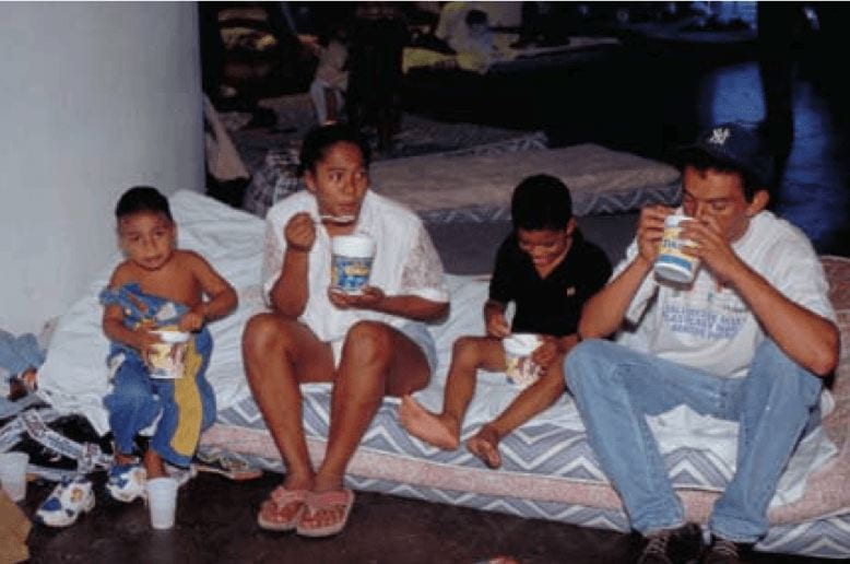 A family of four--parents and two children--sitting on a mattress on the floor of a shelter. There are other mattresses on the floor in the background. Te family eats food out of plastic containers.
