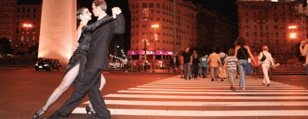 At 3 AM, in the crosswalk in Buenos Aires, a man and a woman dance the tango. Both are wearing formal black attire; she wears a black dress slit up to her upper thigh to show off her leg movement in the dance. Behind them, oridinary people cross the street.