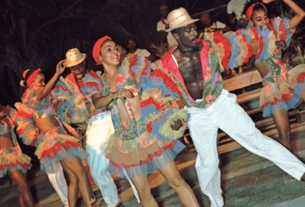 The banrrarra at the villa panamericana in Cojímar, Cuba, east of Havana, performs a Haitian-influenced carnival dance. Men and women dance in highly-colored outfits. The men wear white pants, with a colorful puffy shirt open to the waist. Men and women wear colorful puffy armsleeves.